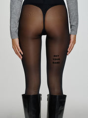 Model trägt schwarze Statement-Strumpfhose mit „NOT YOUR BABE“-Schriftzug auf der Rückseite des Oberschenkels.