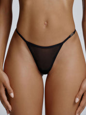 Model trägt den Saint Sass Gigi Mesh Tanga in Schwarz, Vorderansicht, transparenter Mesh-Stoff, vor grauem Hintergrund